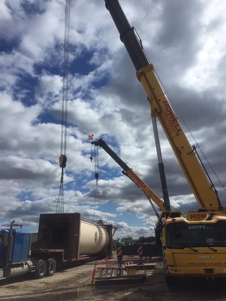 Two yellow cranes lifting a large cylindrical tank from a flatbed trailer against a cloudy sky.