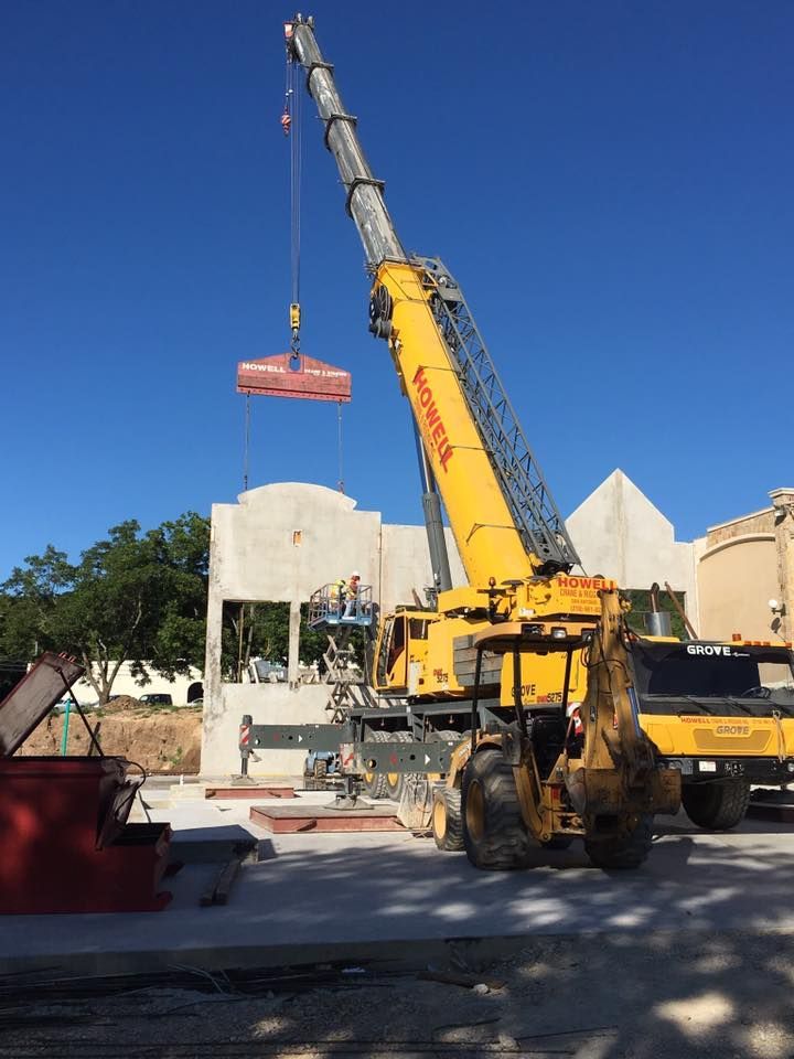 A yellow crane lifts a concrete slab at a construction site. Two workers are visible near a partially built structure under a clear blue sky.