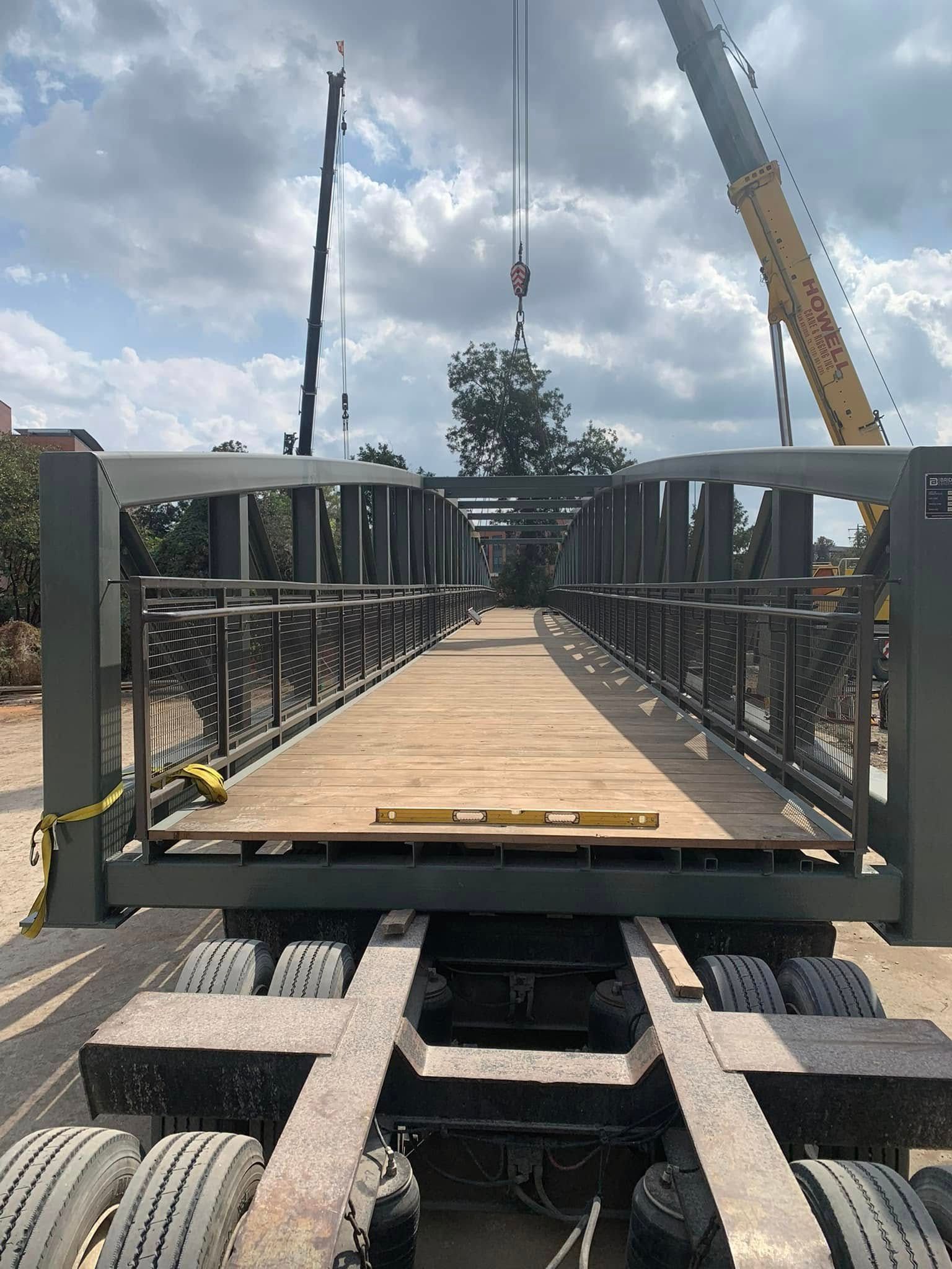 A bridge section being lifted by cranes, positioned over a truck's trailer. The bridge has a wooden deck and metal railings.