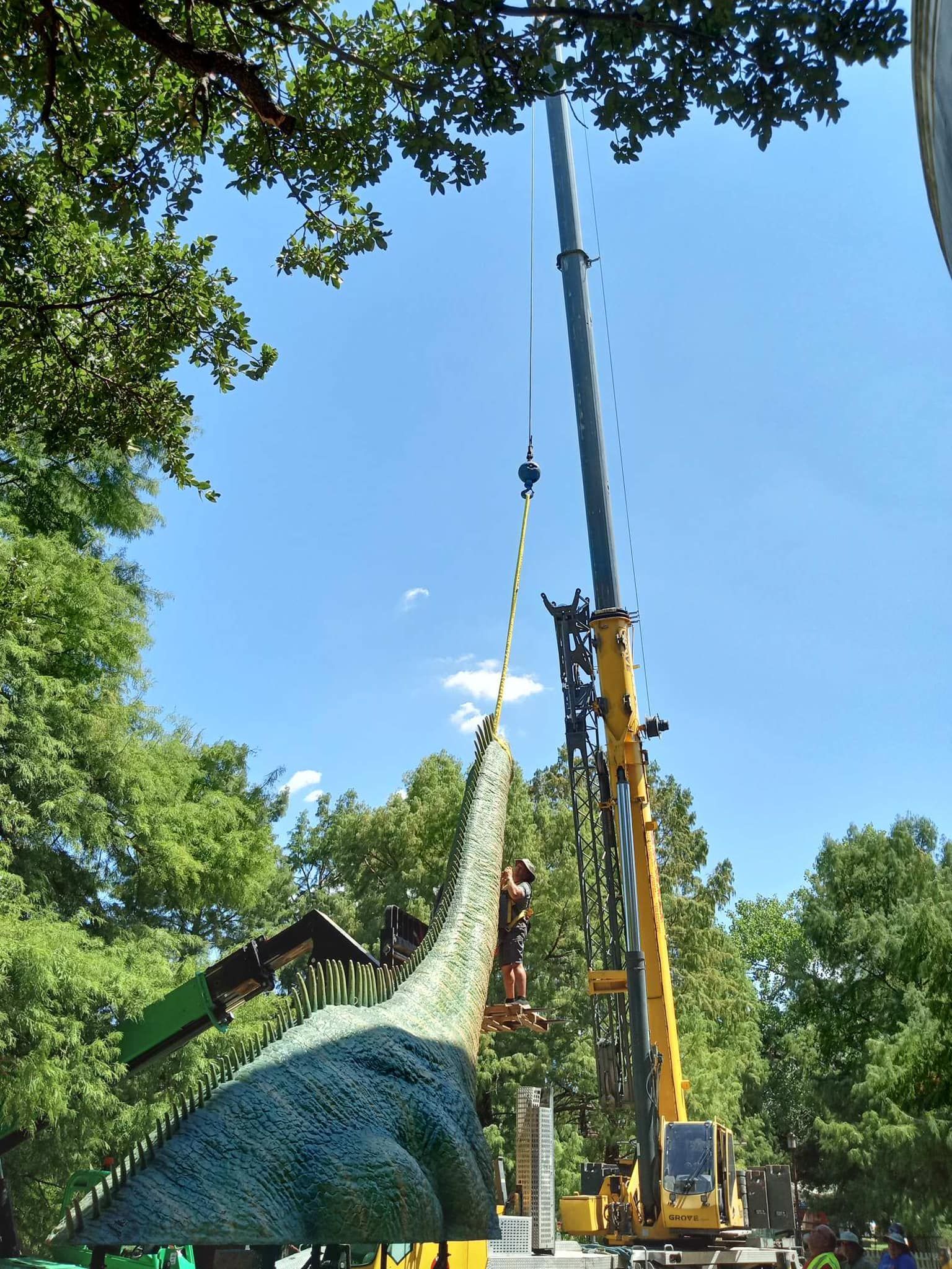 A large yellow crane lifting a green dinosaur statue with trees in the background on a sunny day.