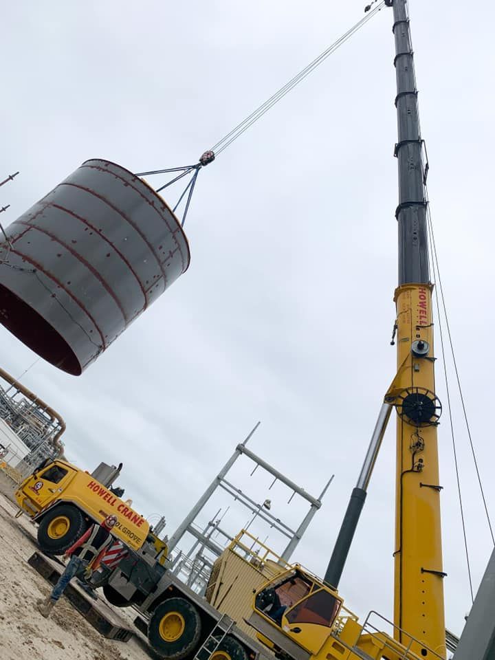 A large yellow crane lifting a cylindrical metal tank at a construction site on an overcast day.