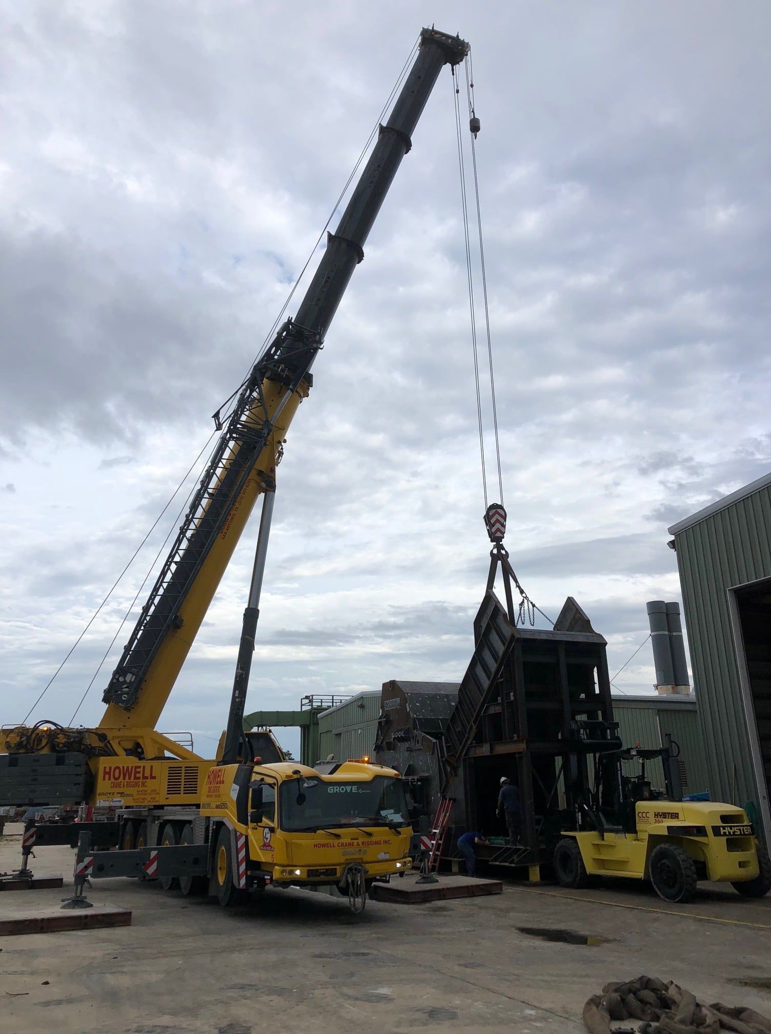 A large yellow crane lifting a metal structure with a forklift assisting on a cloudy day near a building.