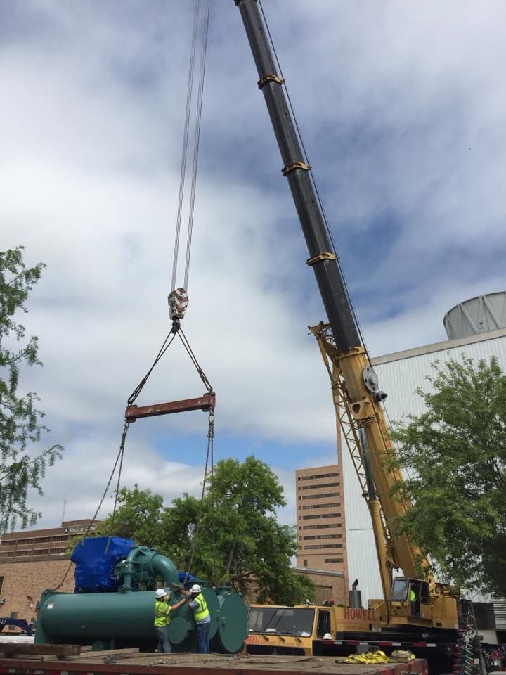 A crane lifts a large green tank with workers nearby in an urban setting, under a cloudy sky.