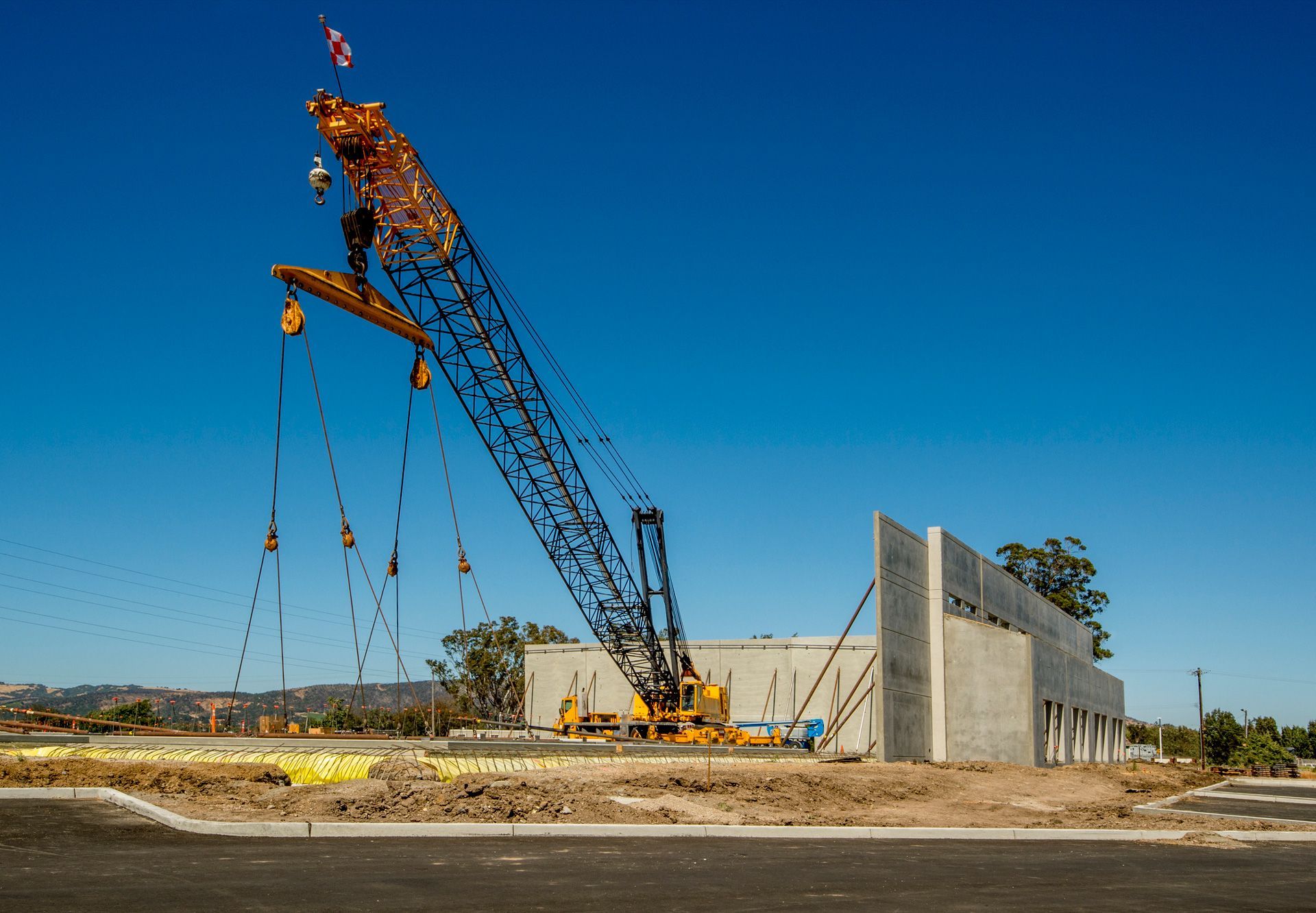 A large crane lifting a concrete wall panel at a construction site on a sunny day. The crane is yellow and black against a clear blue sky.
