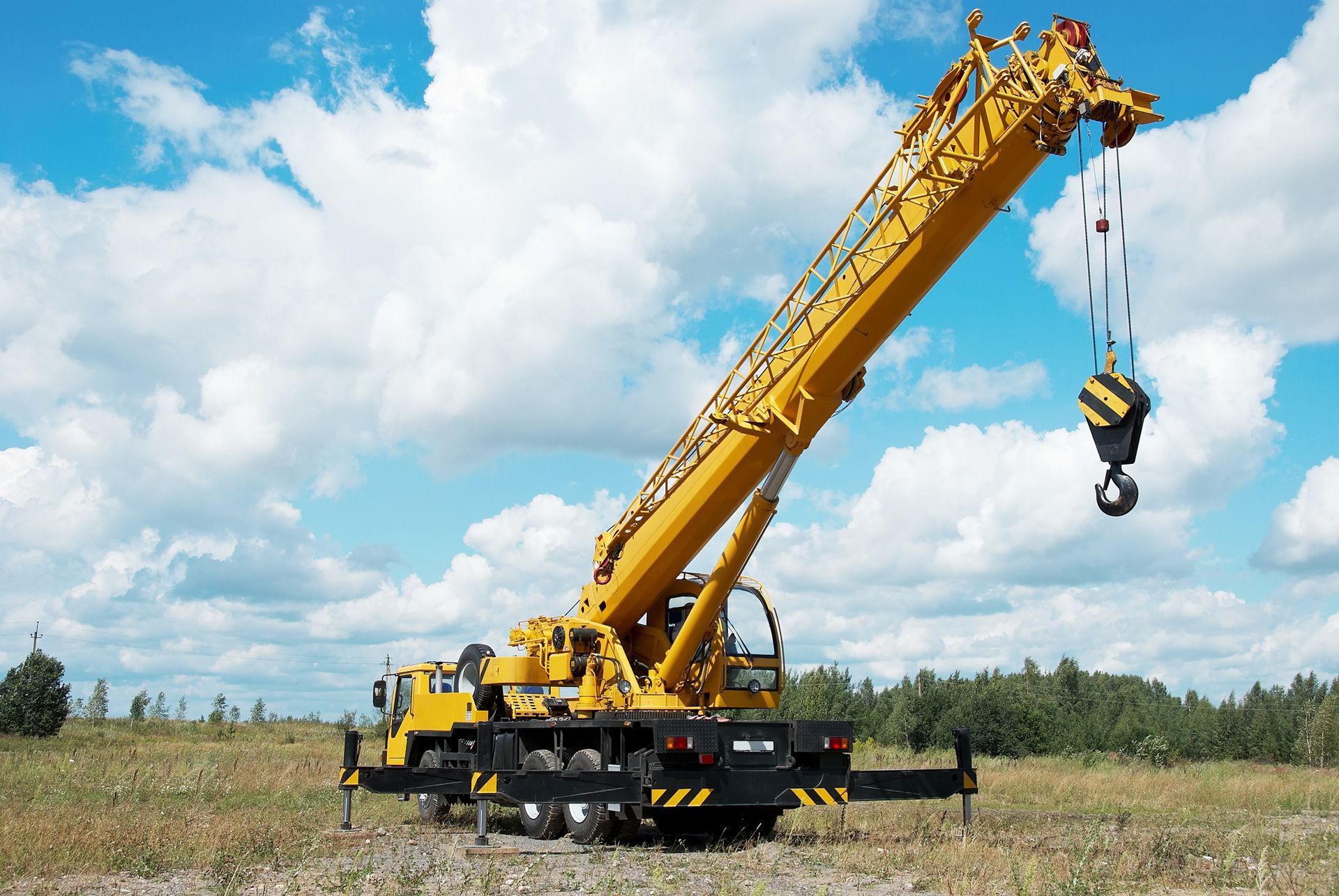 Yellow mobile crane on a sunny day, lifting a heavy object. It is situated in an open field with a blue sky and clouds.