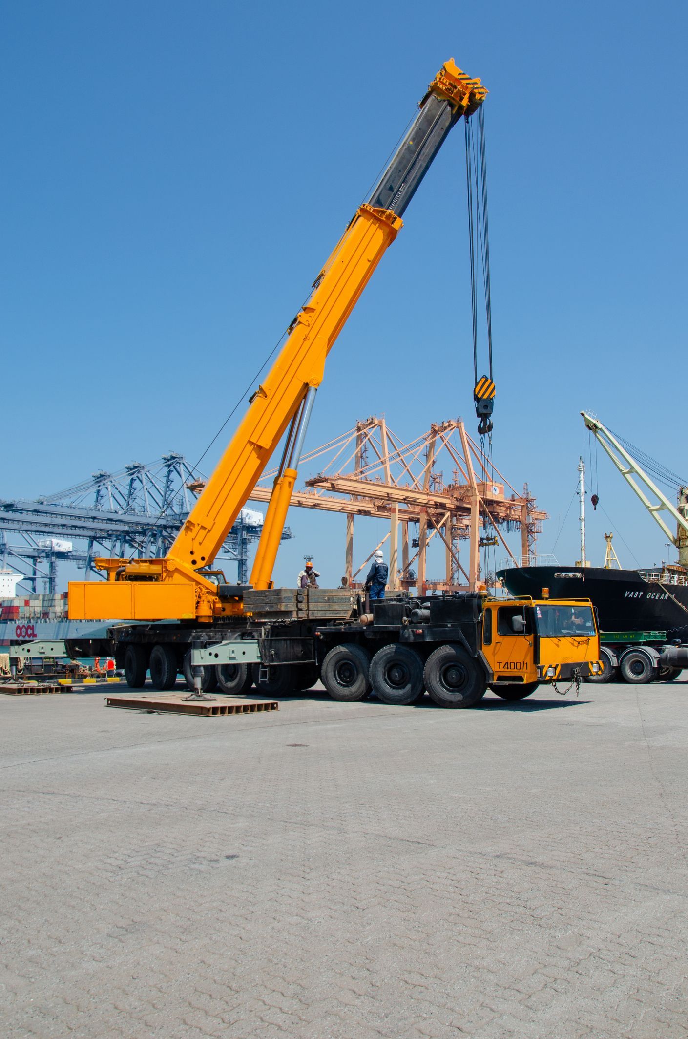 Yellow mobile crane lifting a large metal structure in a harbor setting, with blue sky overhead.