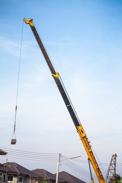 A yellow crane lifting a rectangular object against a light blue sky, near houses and power lines.
