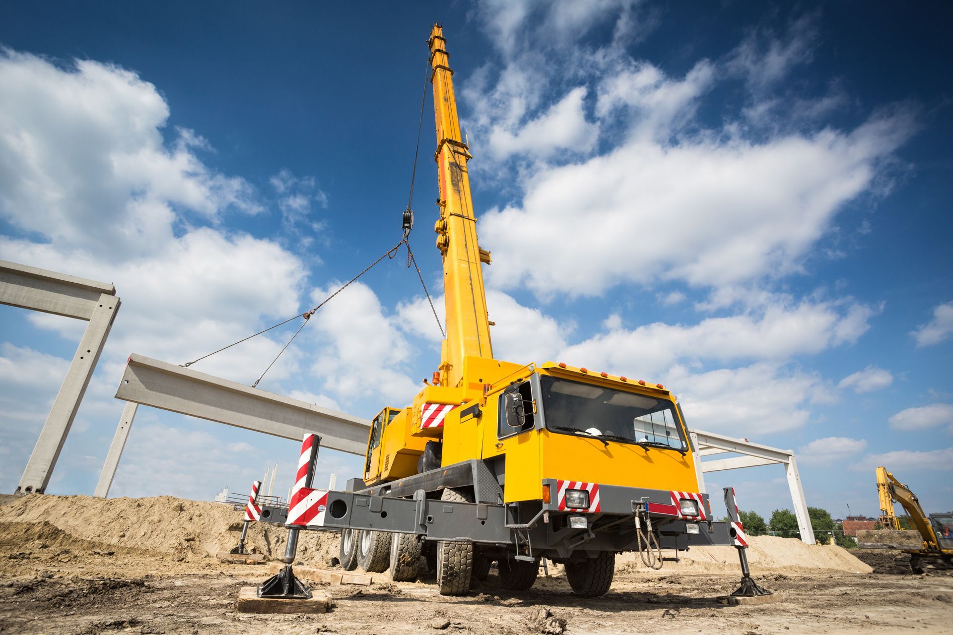 Yellow crane lifting a concrete beam on a construction site under a blue sky with clouds.
