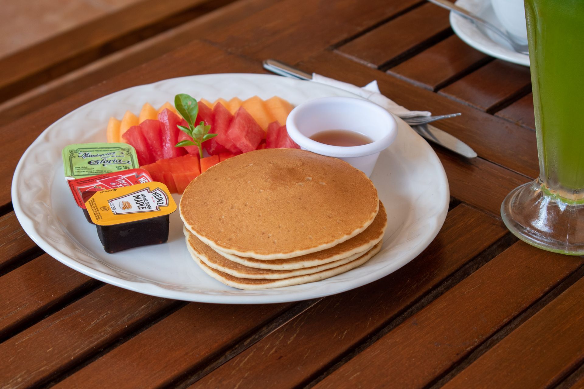 Un plato de panqueques y fruta en una mesa de madera al lado de un vaso de jugo verde.