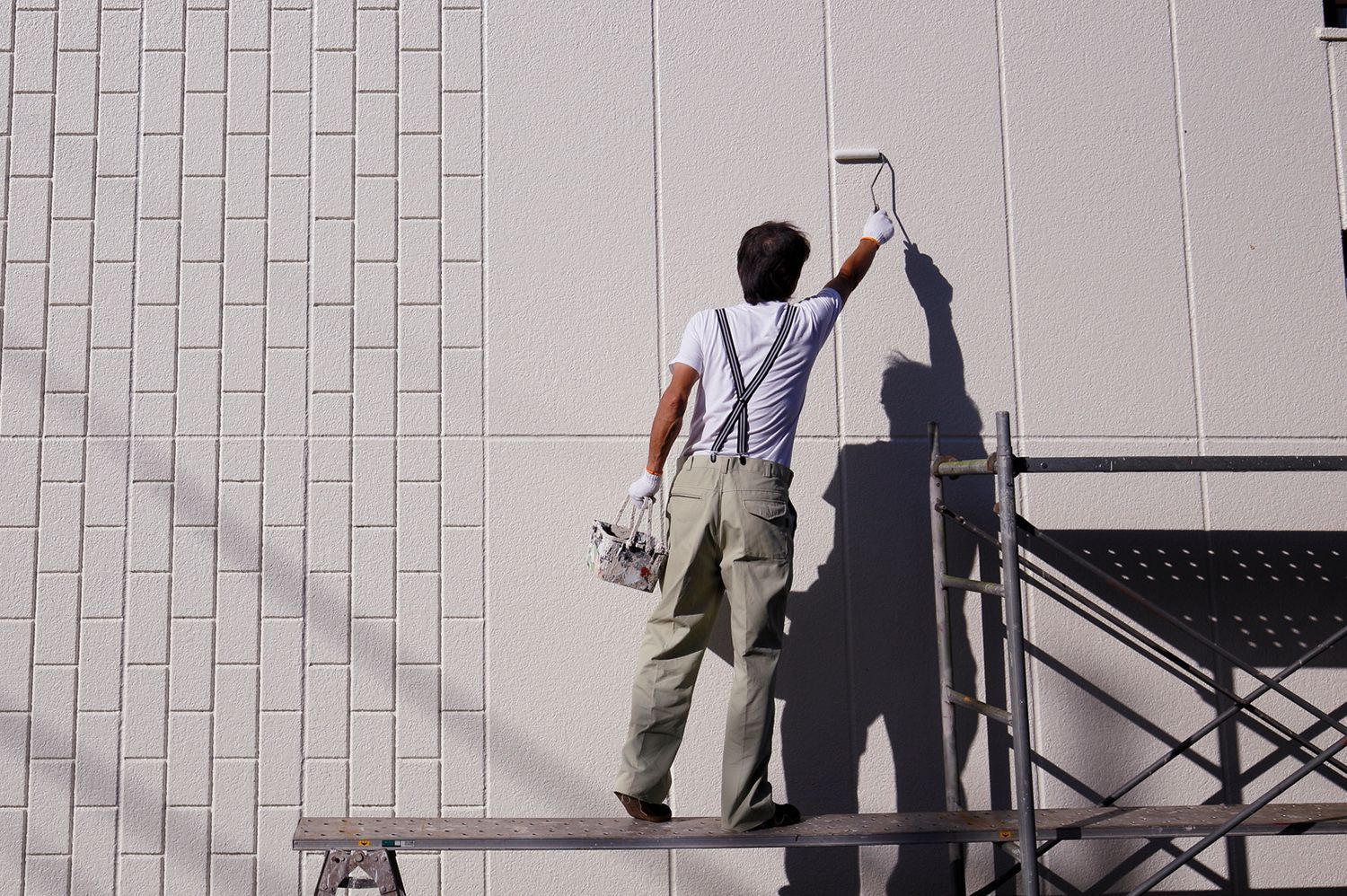 A person paints a light-colored brick wall with a roller, standing on a scaffold outside in the sun. A person paints a light-colored brick wall with a roller, standing on a scaffold outside in the sun.