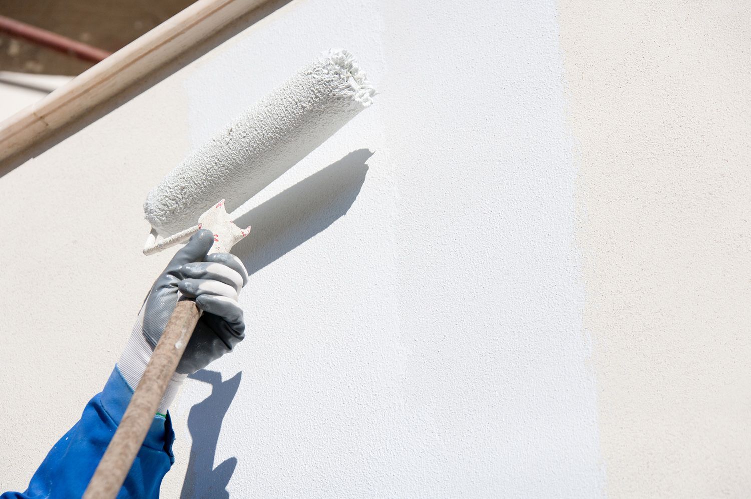 Person painting a white wall with a roller; sunlight creates a shadow.