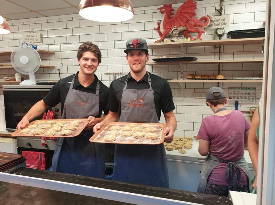 Two men in aprons are holding trays of cookies in a kitchen.
