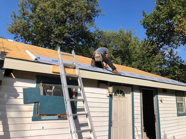 Worker installing shingles on a roof above a white house with a ladder and open doorway below