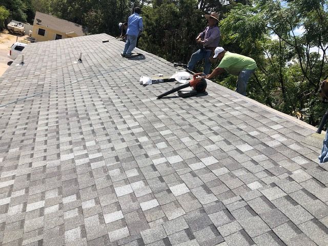 Workers repairing shingles on a steep gray roof near trees
