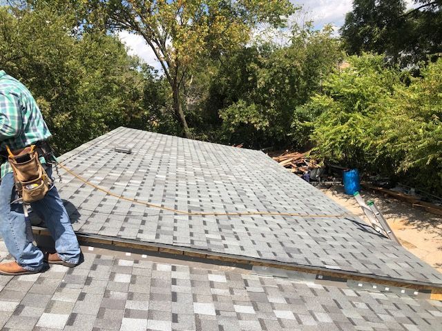 Workers installing gray roof shingles on a house, with trees surrounding the site.
