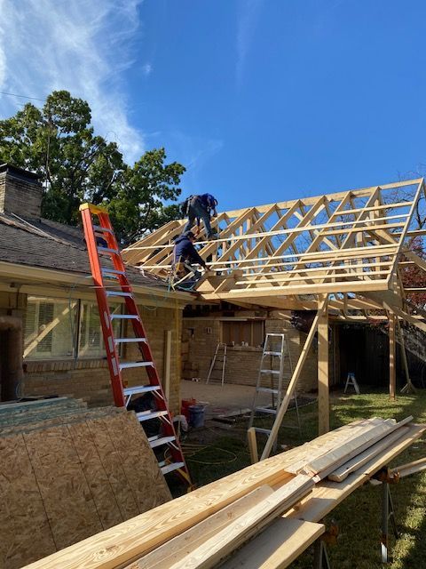 Worker on a roof frame at a house under construction, with ladders and lumber on site.