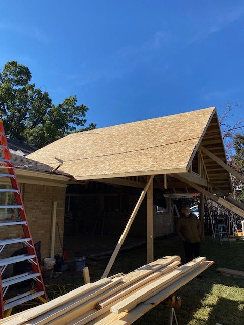 Partially built wooden house with a steep roof frame, lumber in front, and ladders under a blue sky
