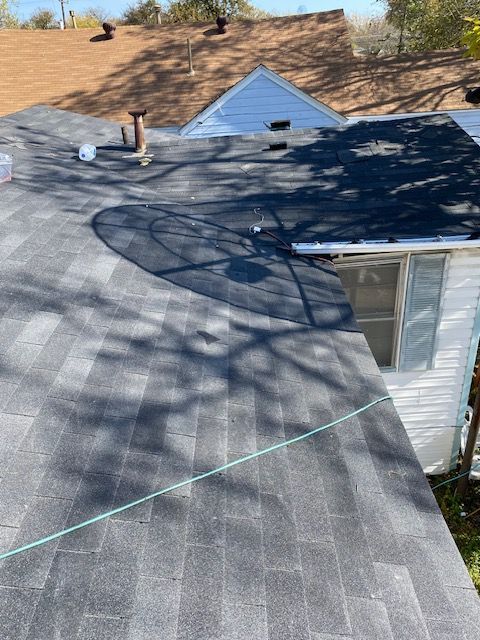 Roofer on a dark shingled roof with safety rope near a white house and blue gable roof in the background
