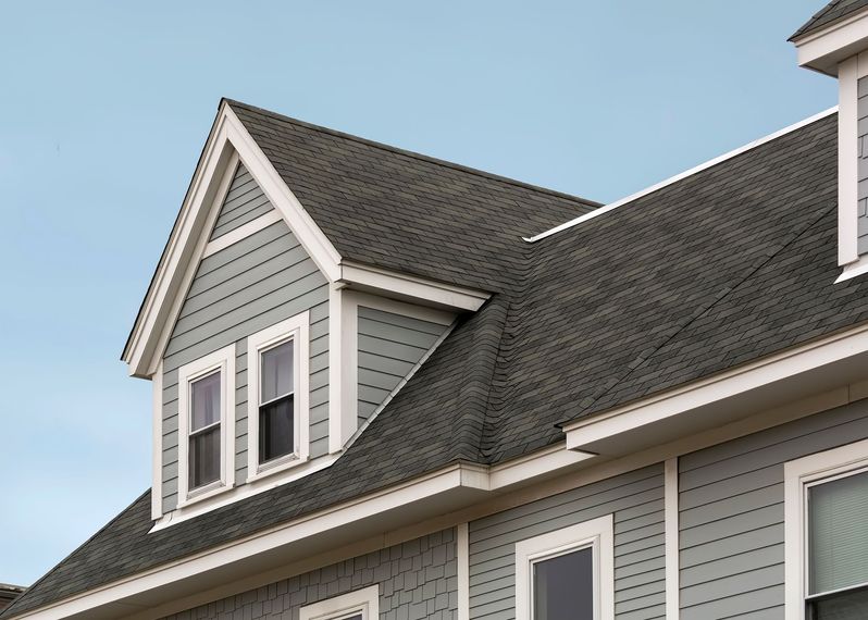 Roofer on a dark shingled roof with safety rope near a white house and blue gable roof in the background