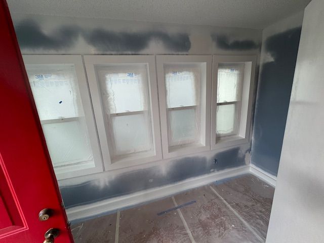 A room with a red door and three windows is being painted.