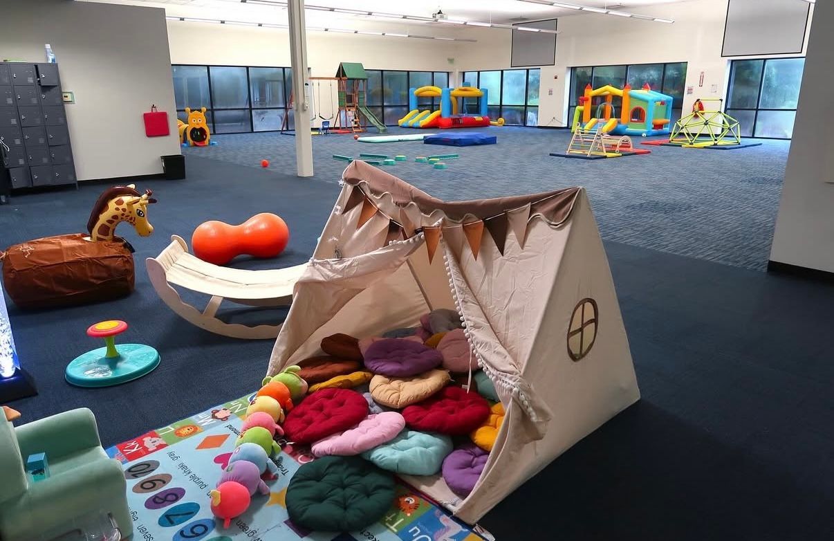 Playroom with tent, toys, and padded flooring; blue carpet, colorful equipment in background.