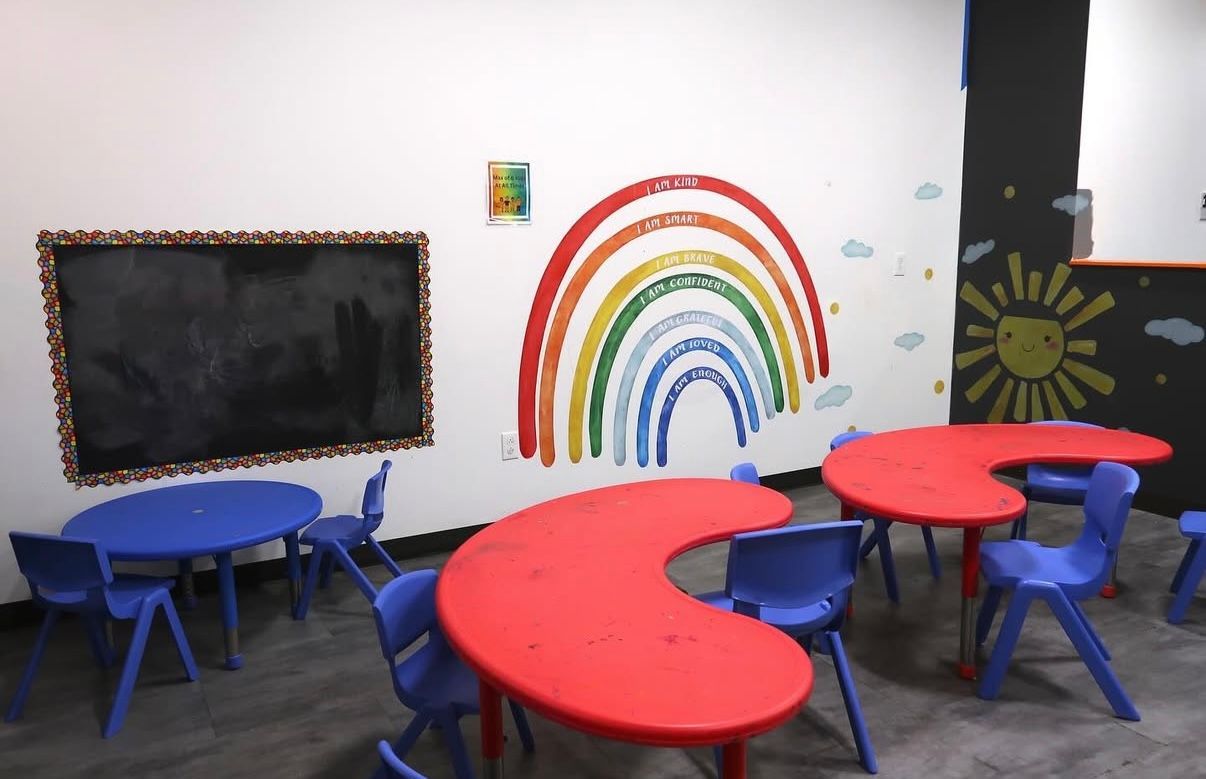 Classroom with colorful rainbow mural, red and blue tables, and chairs.