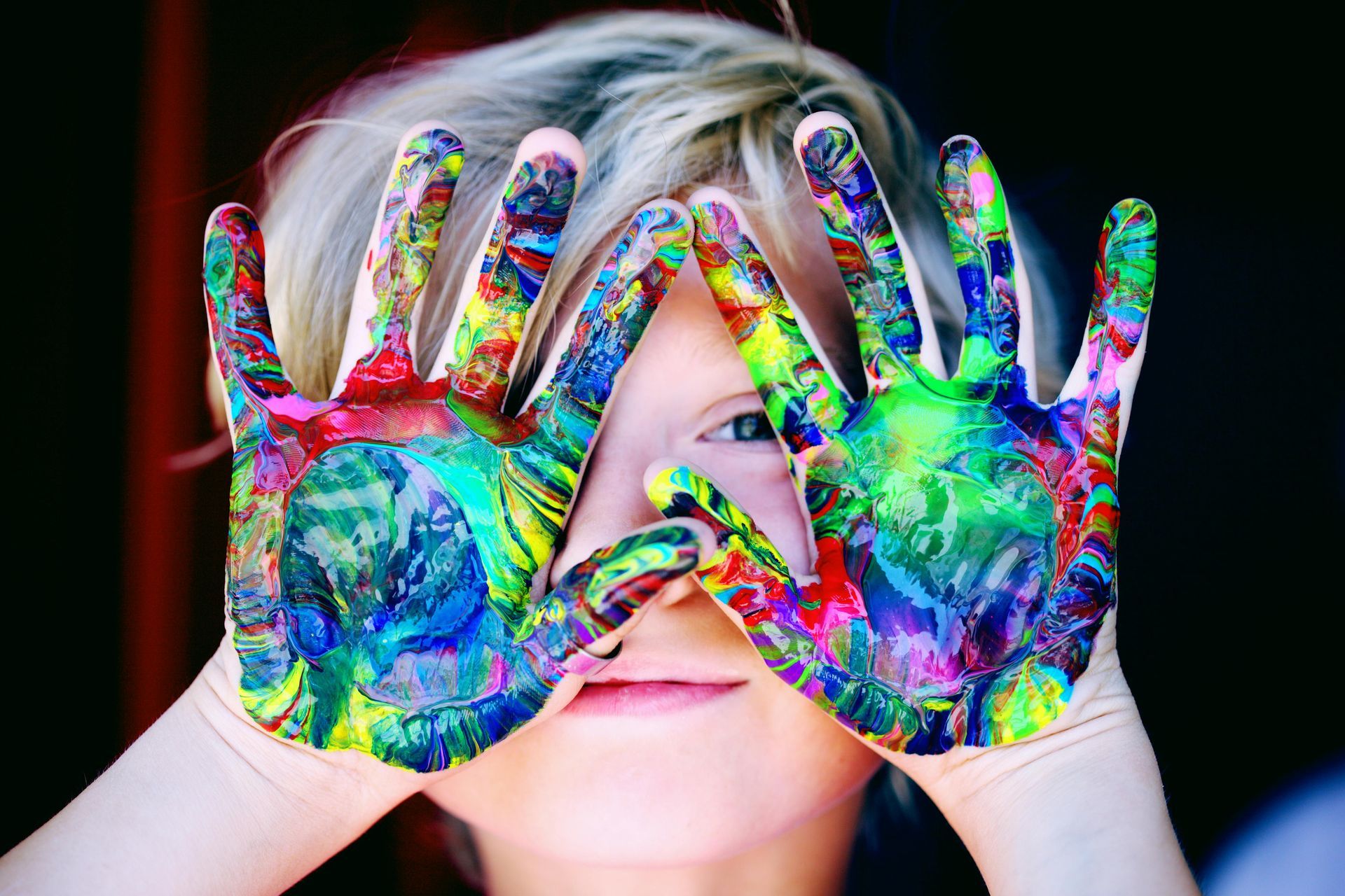 Child looking through hands covered in colorful paint.