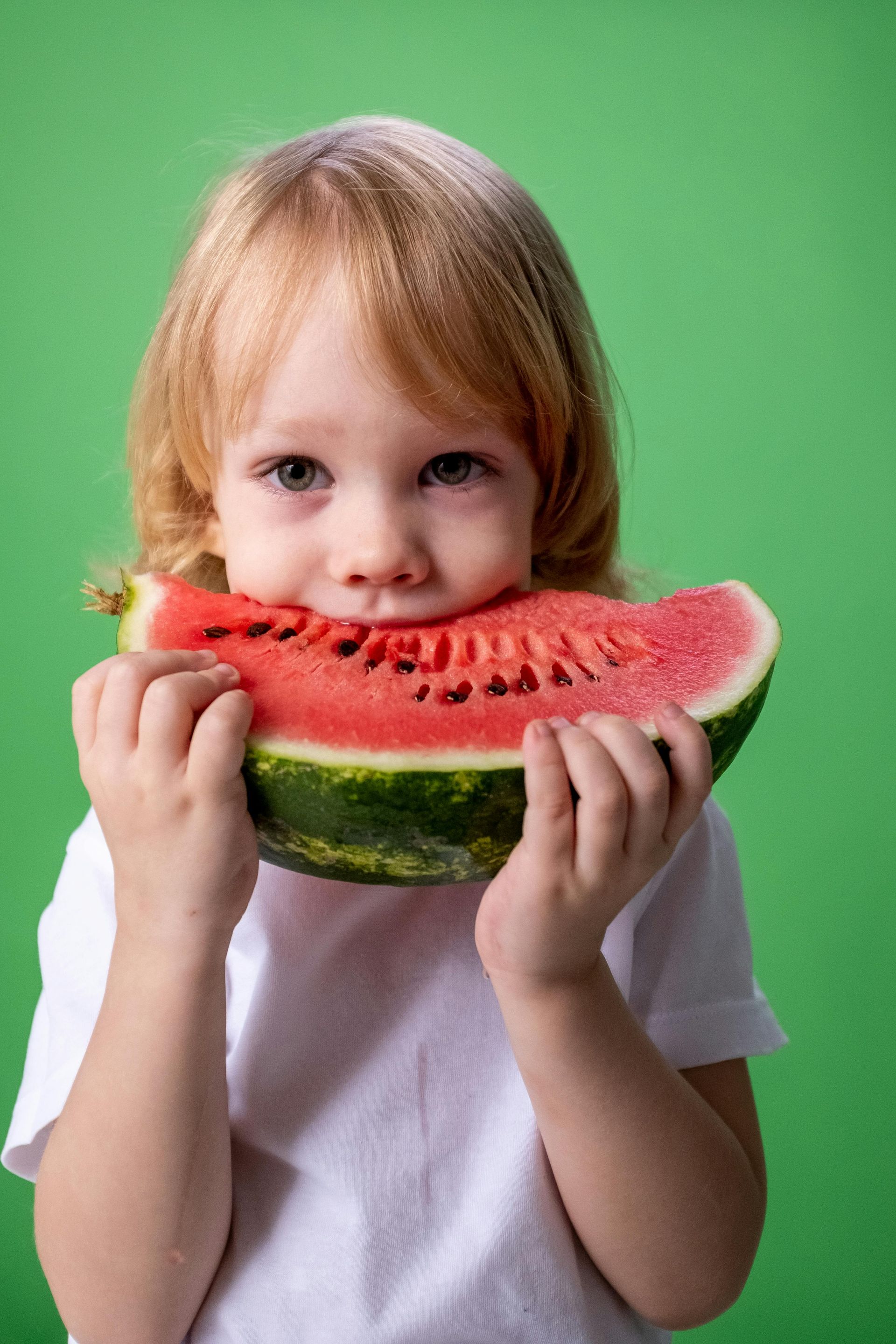Child holding and eating a large slice of watermelon against a green background.