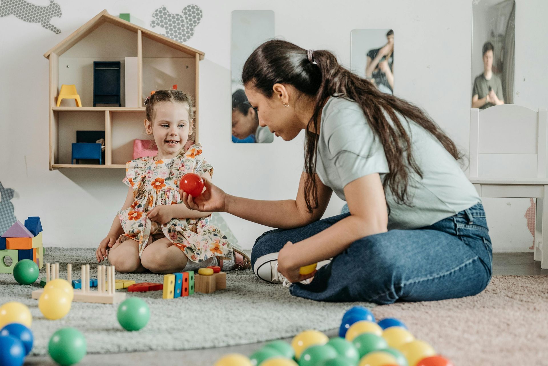 Woman and child playing with colorful balls on a rug in a playroom, surrounded by toys.
