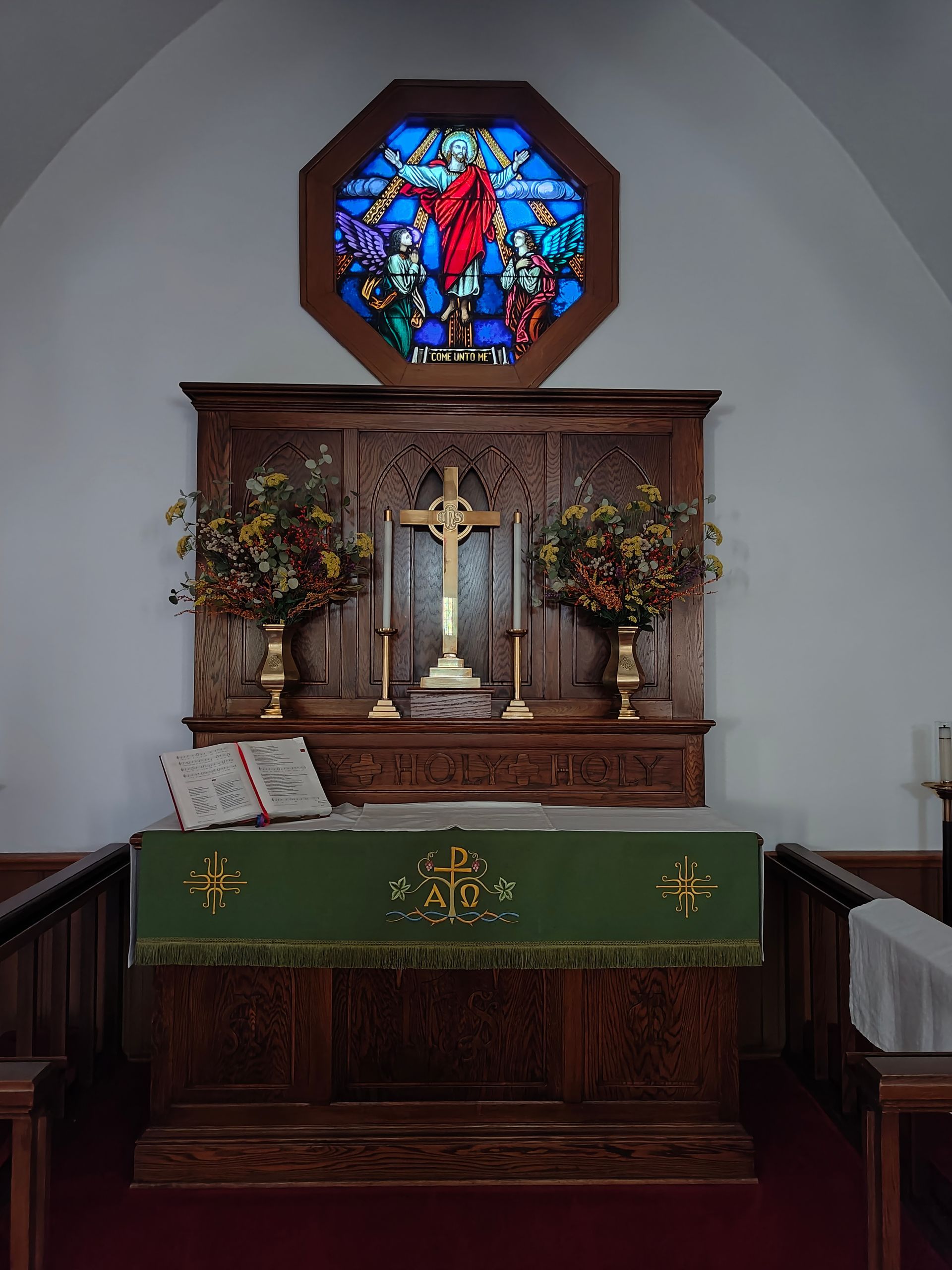 Church altar with stained glass, wooden cross, candles, and floral arrangements.