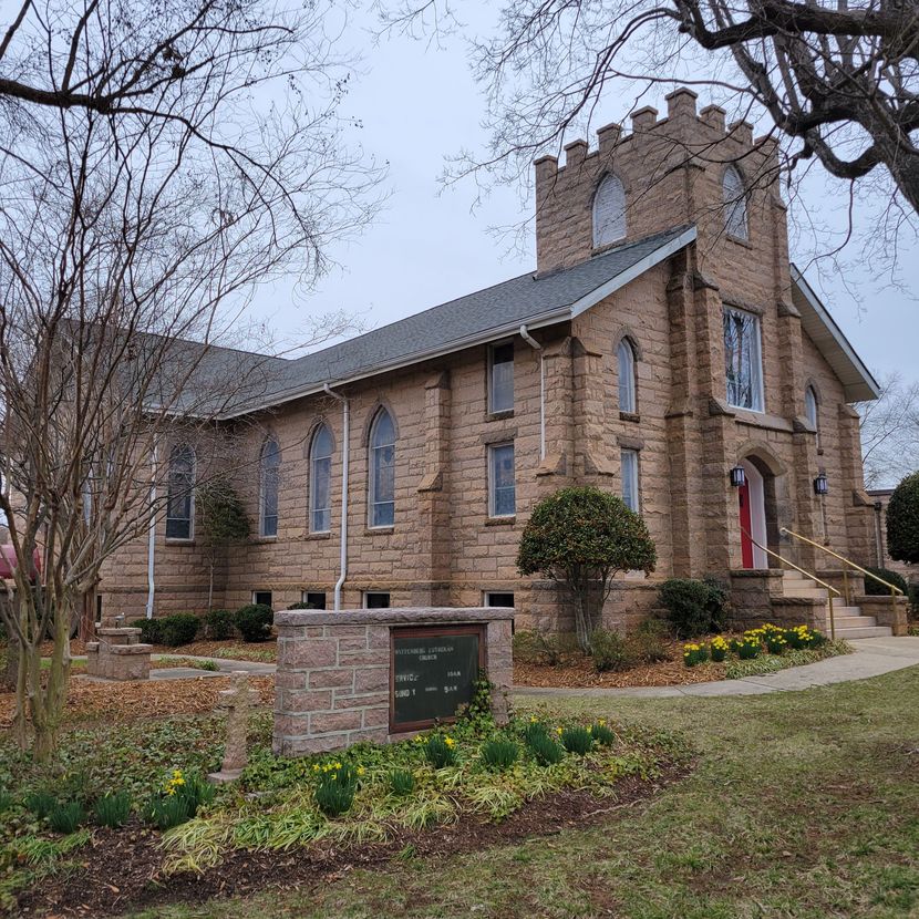 Stone church building with a sign and flowers in the yard.