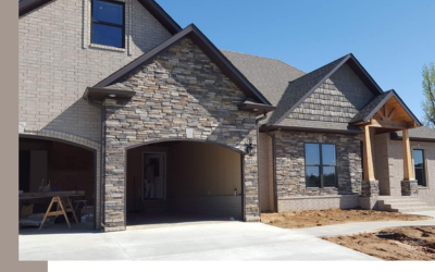 A partially constructed house with gray stone siding, an arched garage entrance, and a front porch with wooden columns.