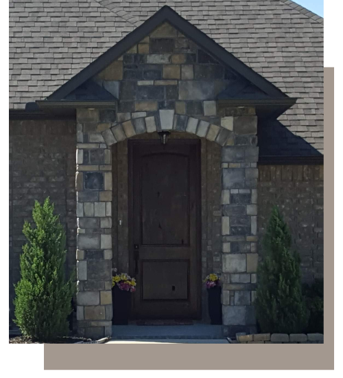 A stone-covered front entryway with a dark wood arched door, flanked by two small evergreen trees and floral planters.