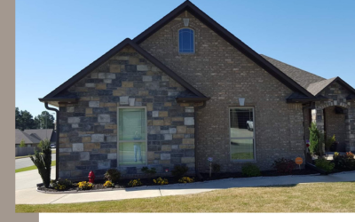 A stone-front residential house with a dark gabled roof and a concrete walkway in front, under a clear blue sky.