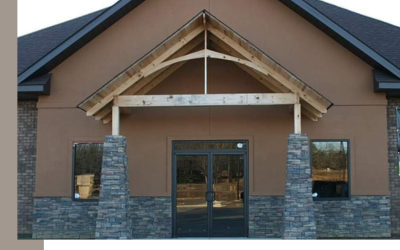 A building entrance featuring a wooden gable roof over double glass doors, flanked by stone columns and tan stucco walls.