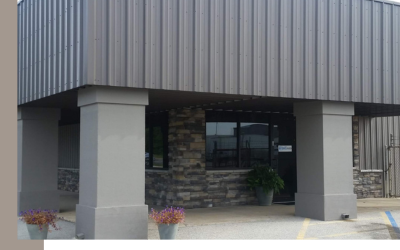 A building entrance with two gray pillars, dark gray metal siding, and stone accents, featuring potted flowers in front.