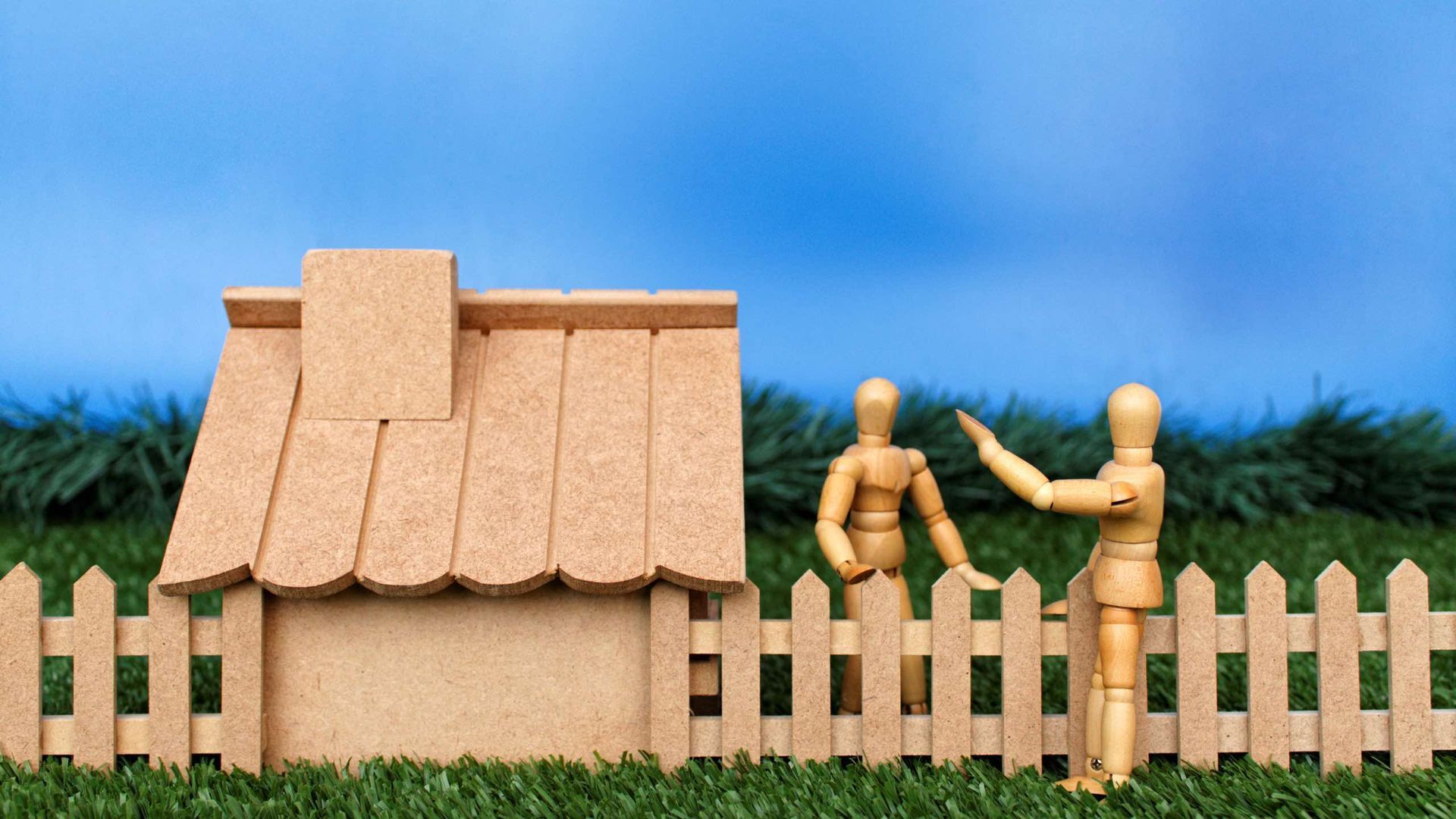 Two wooden mannequins are standing next to a wooden fence and a wooden house.