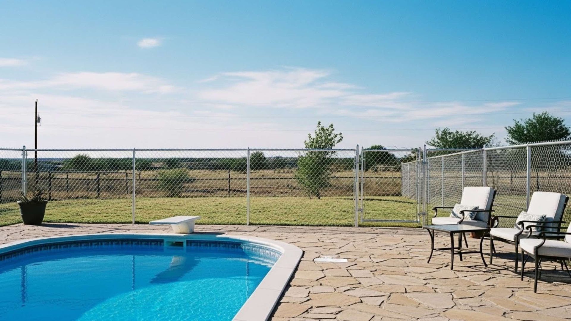Oval pool with diving board, patio, chairs, and chain-link fence on a sunny day.