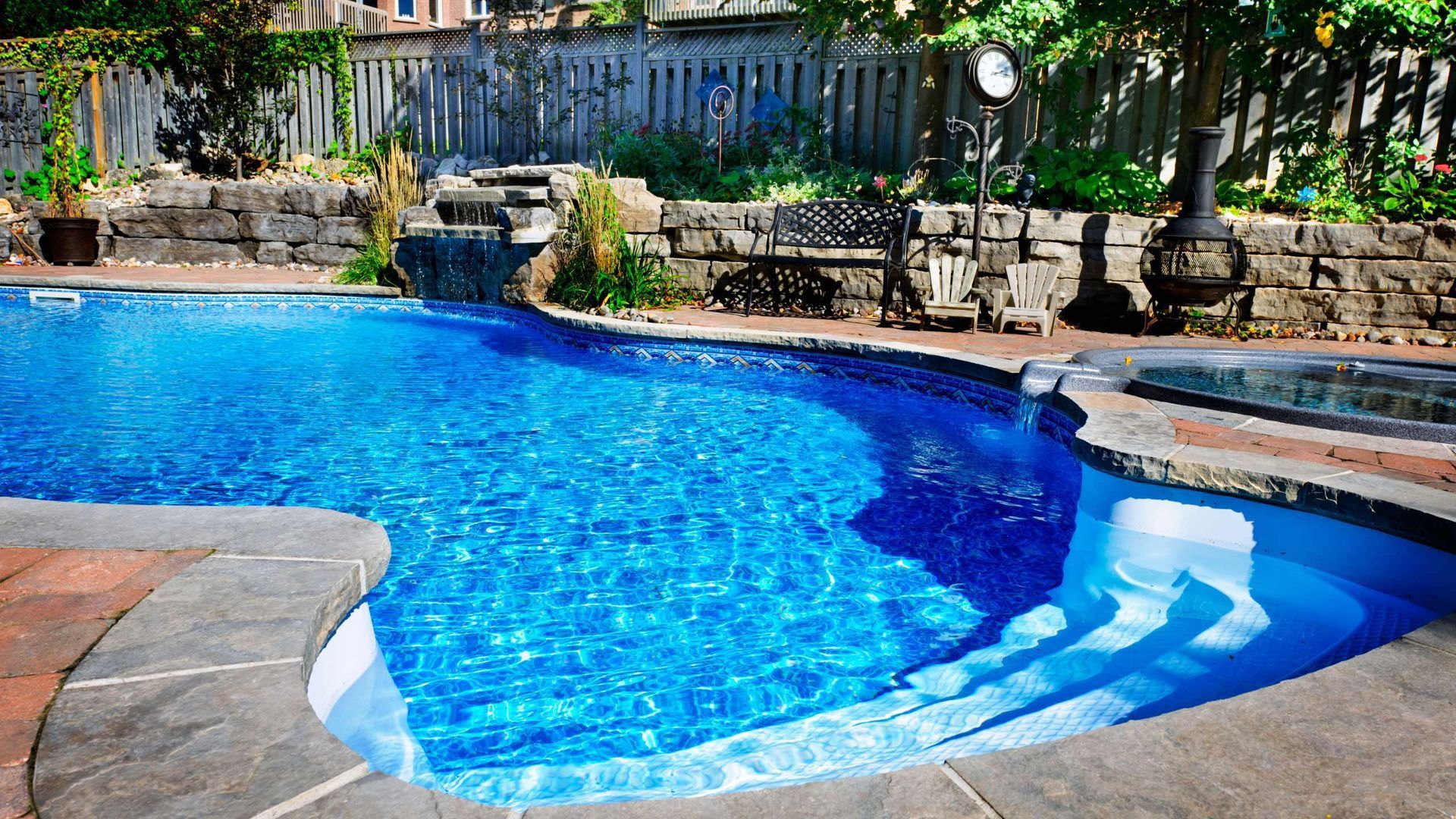 Blue swimming pool with stone steps and patio, surrounded by stone walls and greenery.