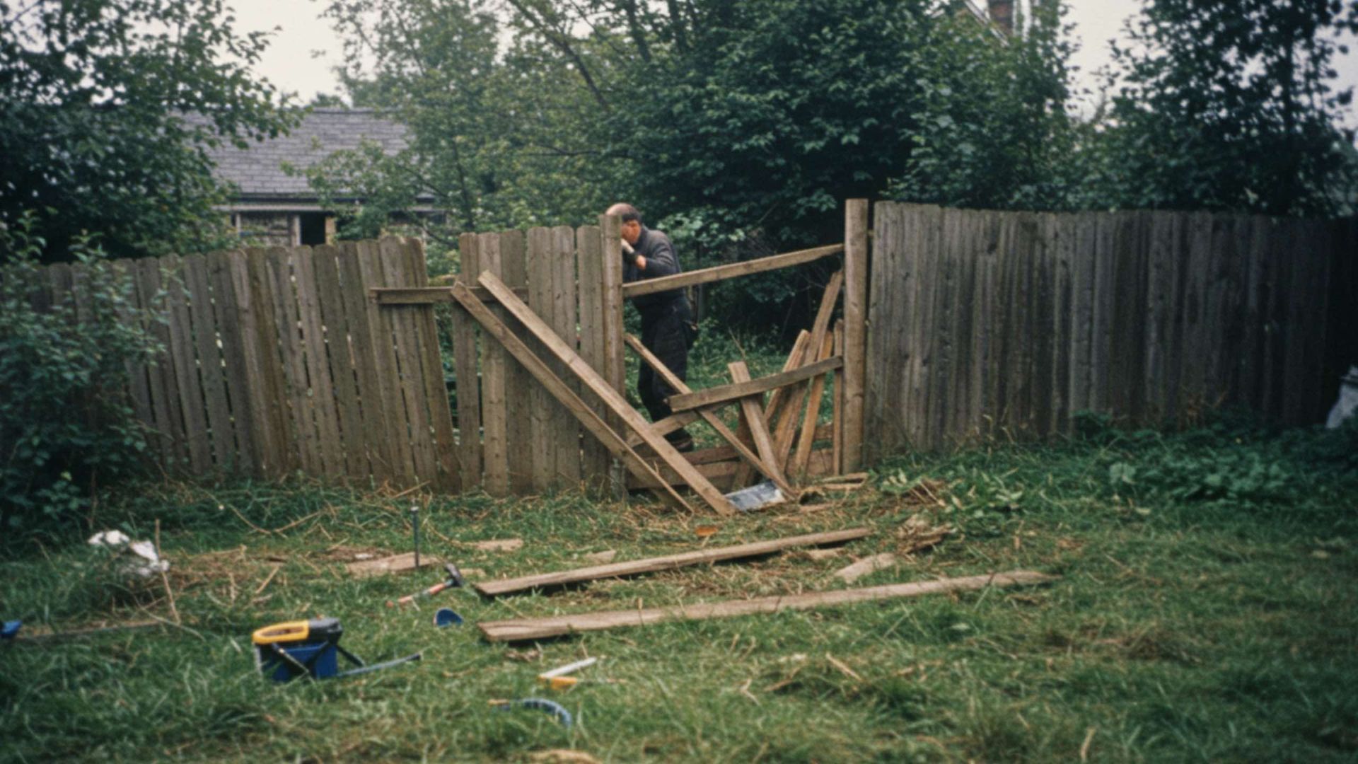 Man repairing a broken wooden fence in a grassy yard, tools scattered around.