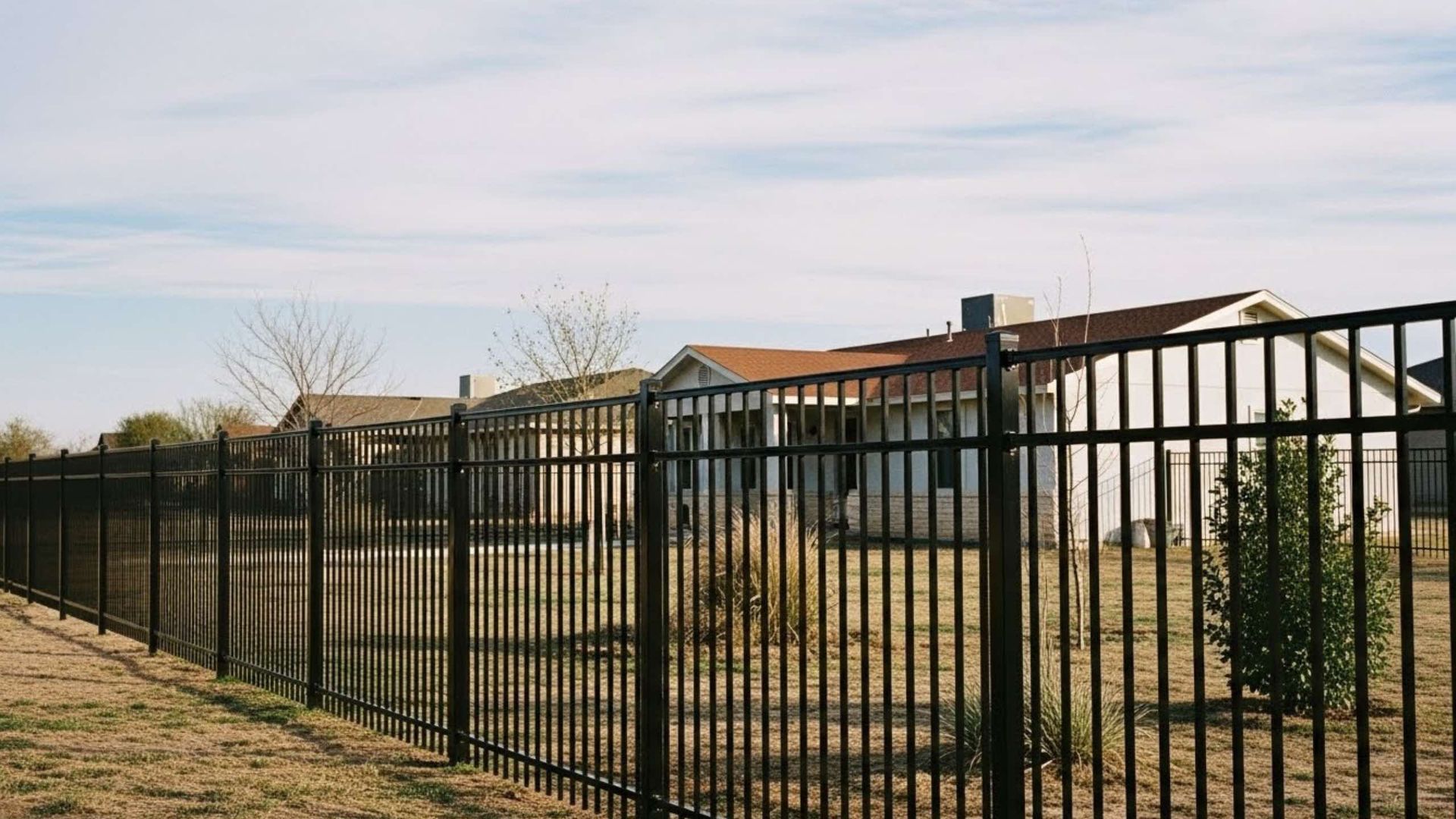 Black metal fence in front of several houses with brown roofs under a blue sky.