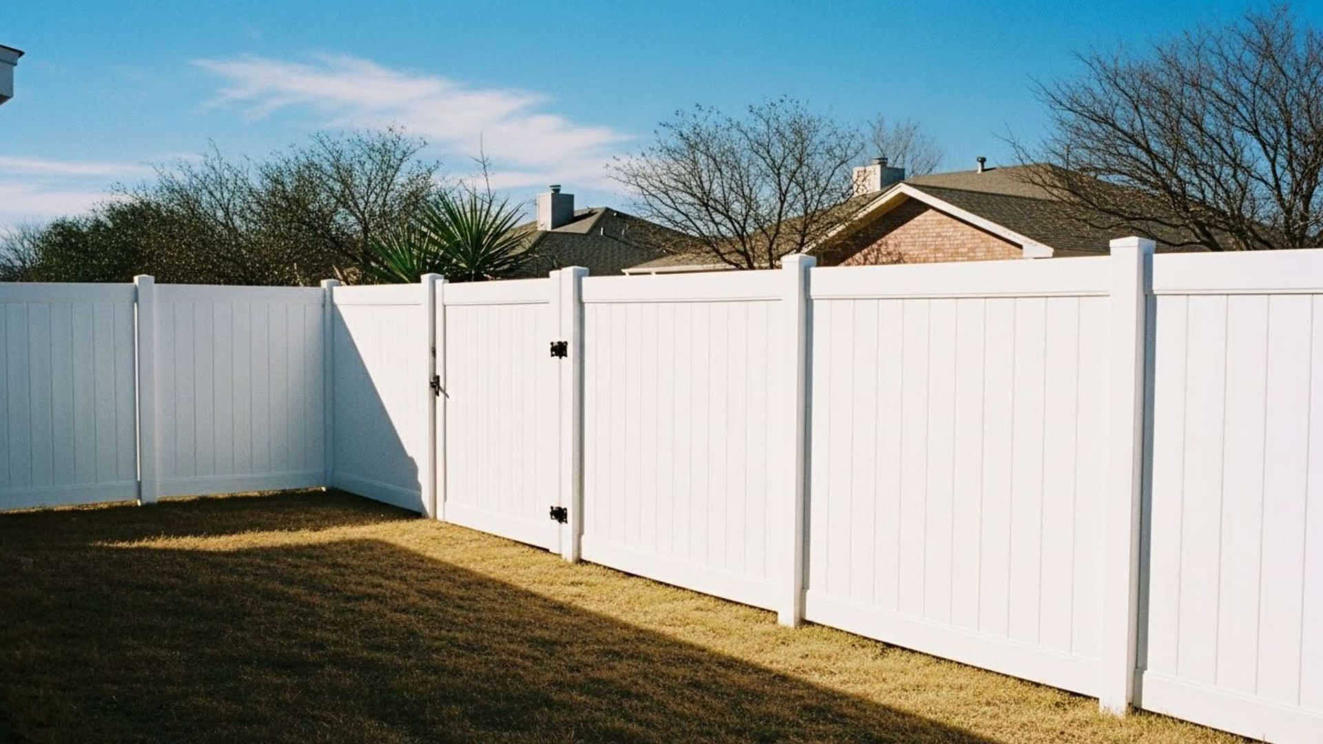 A white vinyl privacy fence surrounds a backyard with dry grass under a blue sky.
