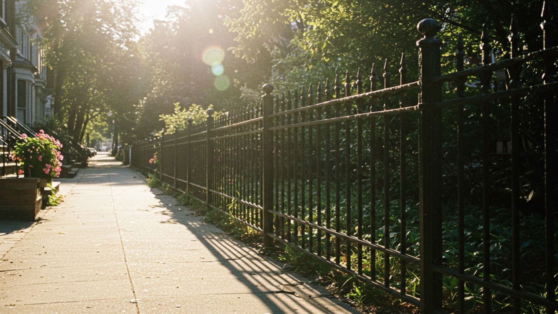 Sunlit sidewalk with wrought-iron fence and trees, casting shadows. Houses on the left.