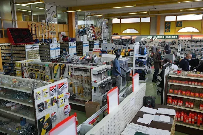 Intérieur d'une quincaillerie rempli d'outils, d'étagères et de clients. Murs jaunes, présentoirs blancs.
