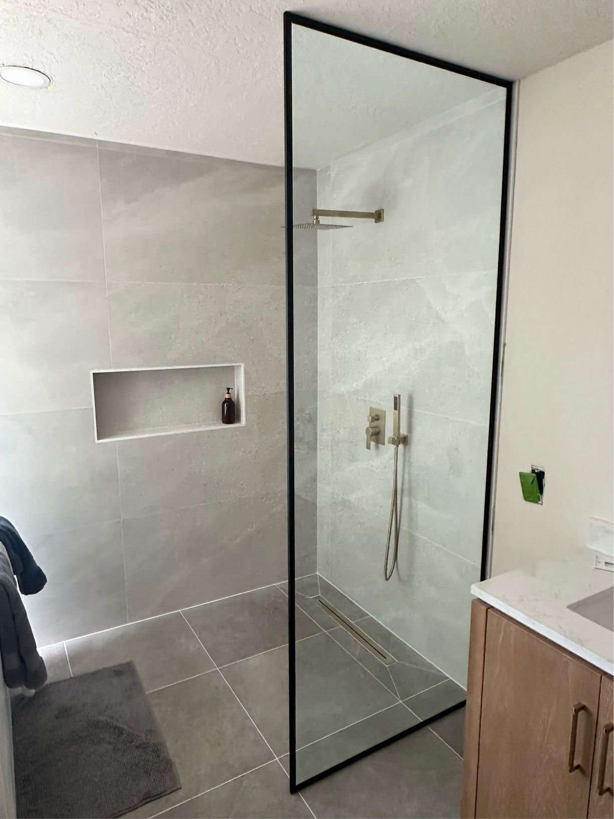 Shower with glass enclosure and black frame, grey tile, and wooden vanity.