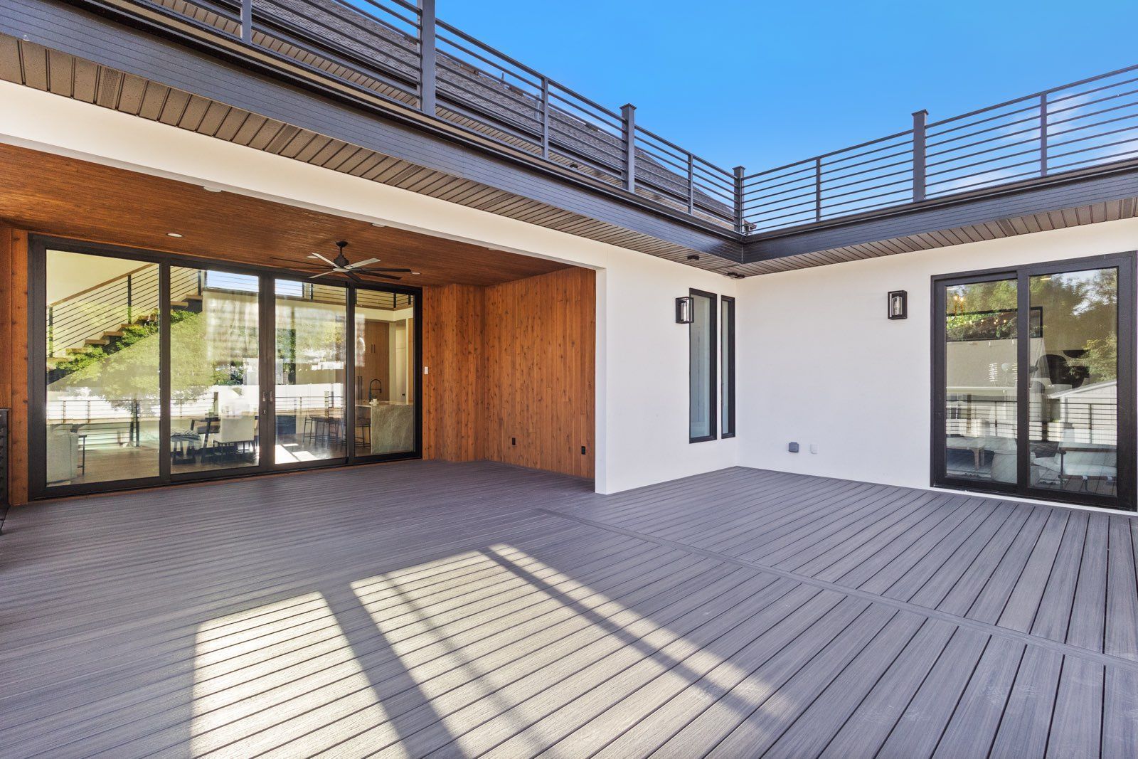 Spacious outdoor patio with wooden deck, sliding glass doors, and rooftop deck under a blue sky.