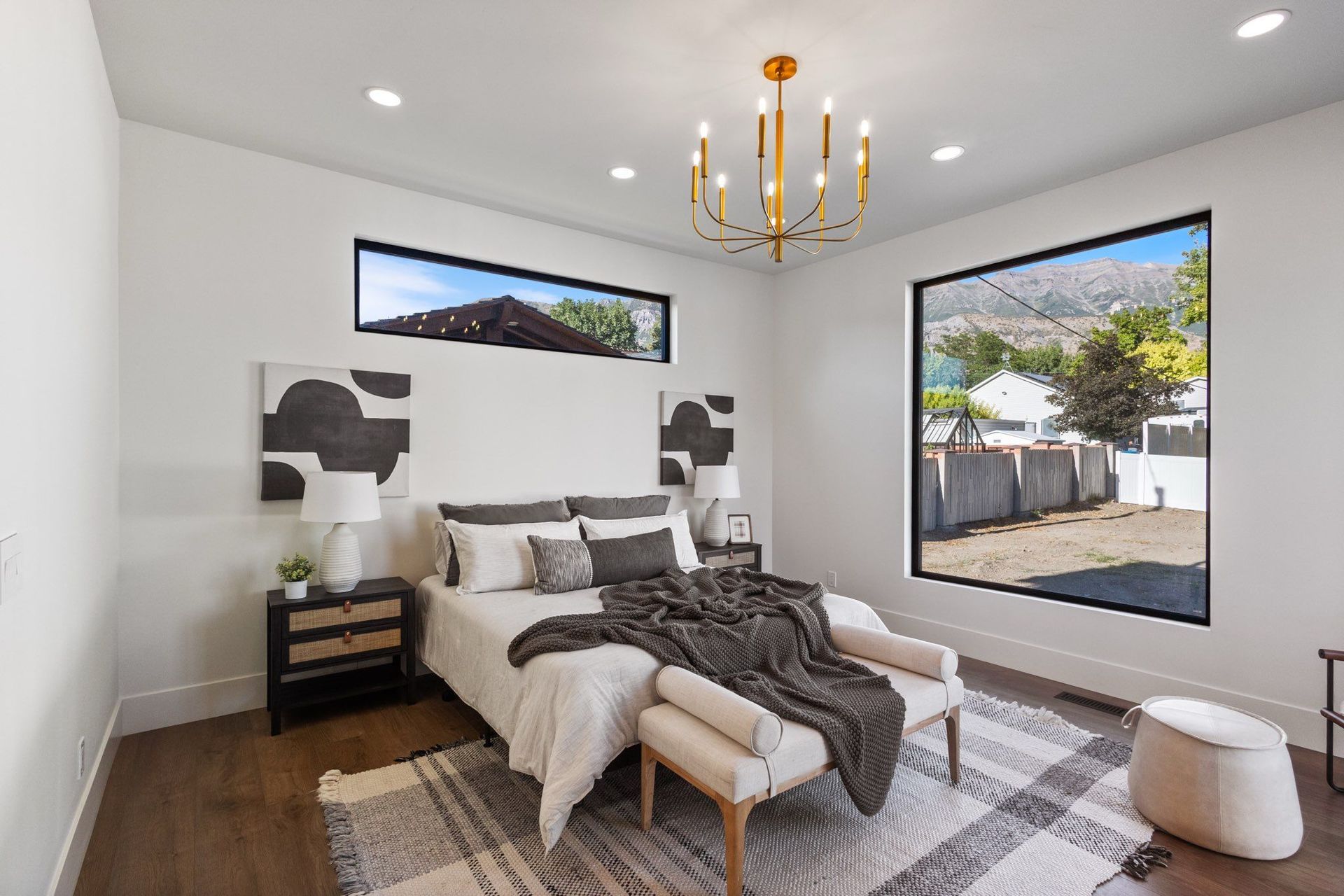 Modern bedroom with bed, two nightstands, large windows, and a gold chandelier.
