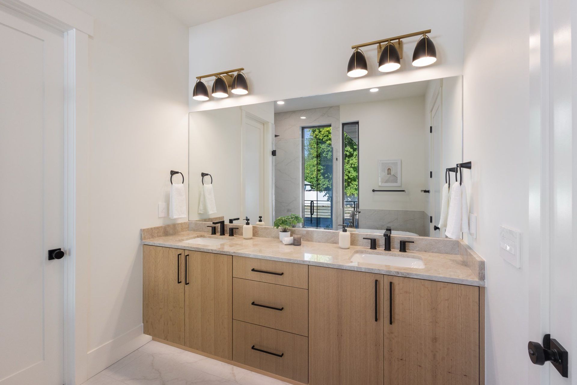 Bathroom with double vanity, light wood cabinets, and large mirror. Brass light fixtures, black hardware, white walls.