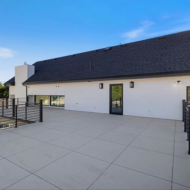 Exterior of a modern home with a concrete patio. White walls, black roof, glass door. Sunny, blue sky.
