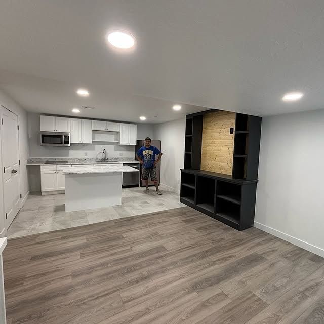 A person stands in a newly renovated basement with a kitchen, island, built-in shelving, and wood-look flooring.
